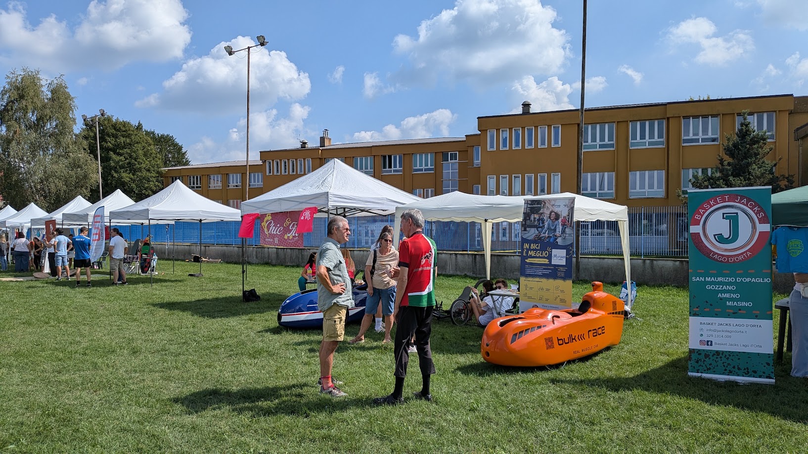 Vista ampia dello stand dell’ASD Velocità a Pedali, con un velomobile blu e uno arancione sul prato